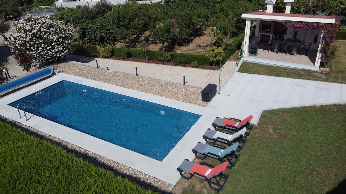 An inviting outdoor area showcases a large pool surrounded by a stone patio. Colorful lounge chairs are arranged along the poolside. A pergola with seating can be seen nearby, nestled among lush greenery, providing a relaxing space for guests to unwind.