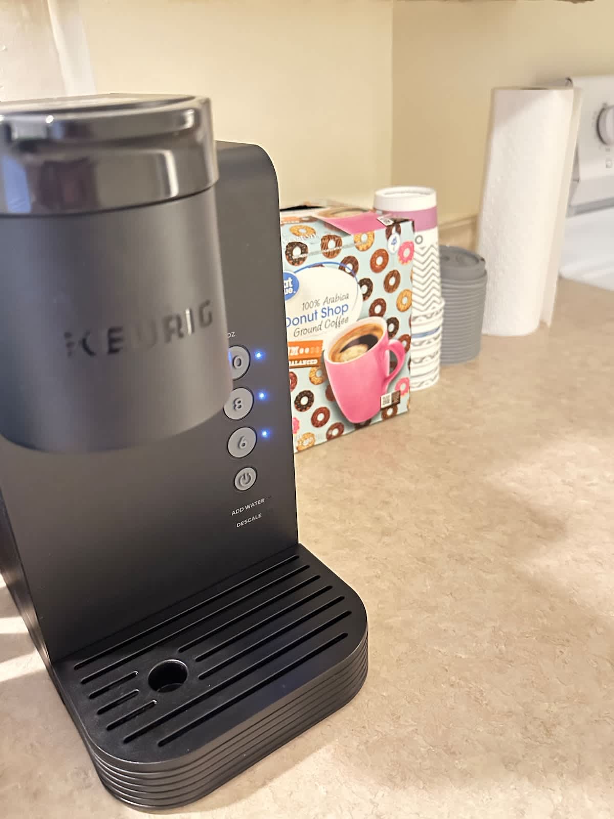 A Keurig coffee maker is positioned on a beige countertop, featuring a sleek black design with blue buttons. Behind it, a box of coffee pods from The Donut Shop is visible, alongside disposable cups and paper towels, providing convenient options for beverage preparation.