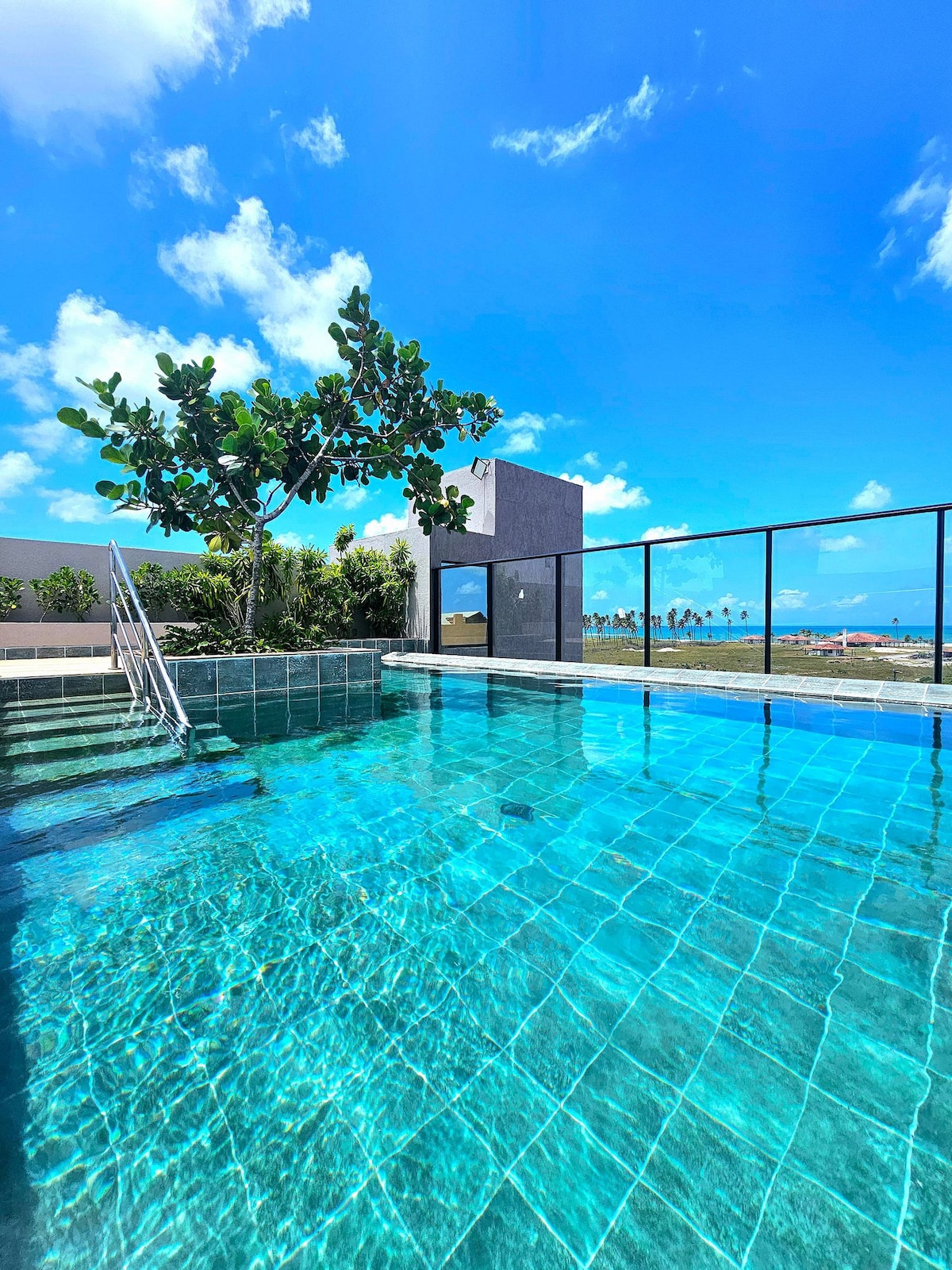 A clear blue pool glistens under the bright sky, with steps leading into the water. Lush greenery is visible nearby, alongside a modern structure in the background. The expansive view showcases coastline and ocean, enhancing the outdoor relaxation space.