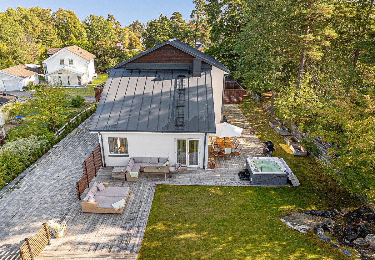 An aerial view of a spacious outdoor area surrounding a modern house. A private jacuzzi is located next to a wooden deck. Comfortable seating is arranged on the deck, with dining furniture visible for outdoor gatherings. Lush greenery and trees frame the scene.
