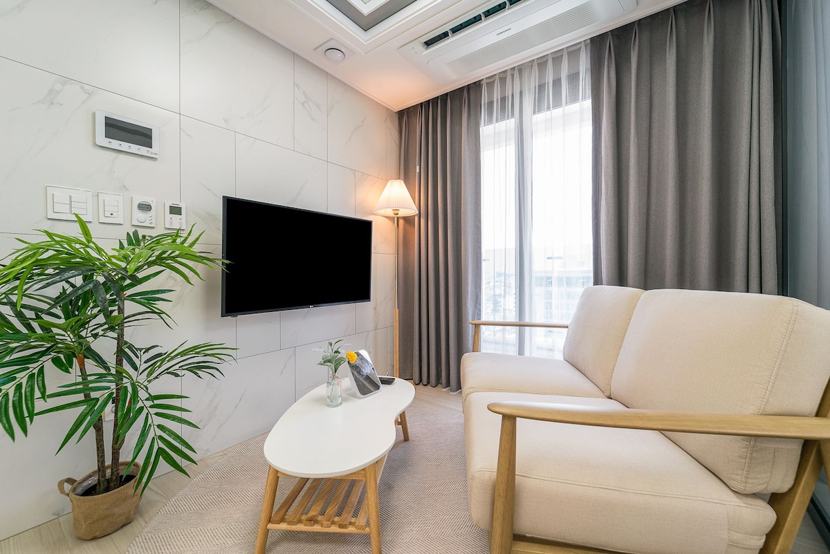 A bright living area features a light-colored sofa placed next to a stylish coffee table. A flat-screen TV is mounted on the marble wall, with a small potted plant adding a touch of greenery. Natural light filters through sheer curtains, enhancing the ambiance.