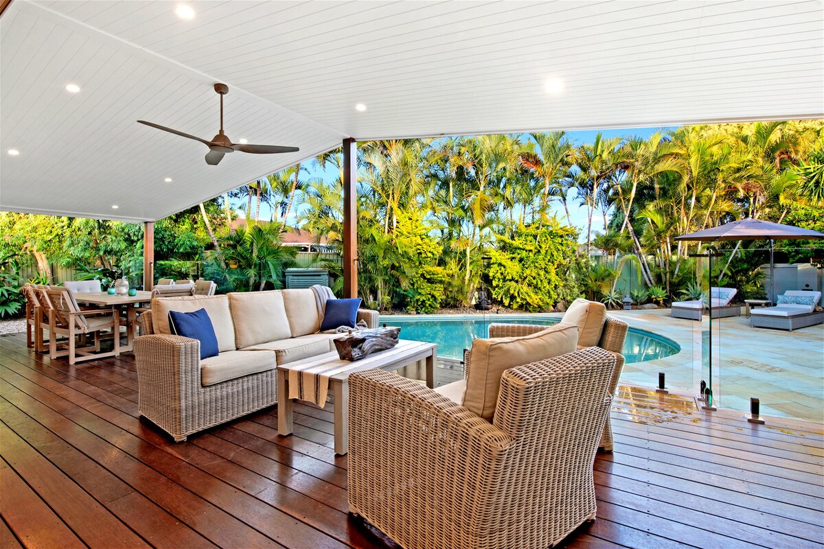 A spacious outdoor living area features stylish wicker furniture arranged around a low table, overlooking a heated swimming pool. Lush tropical landscaping surrounds the space, providing a sense of privacy. A dining table is positioned nearby, creating an inviting atmosphere for gatherings.