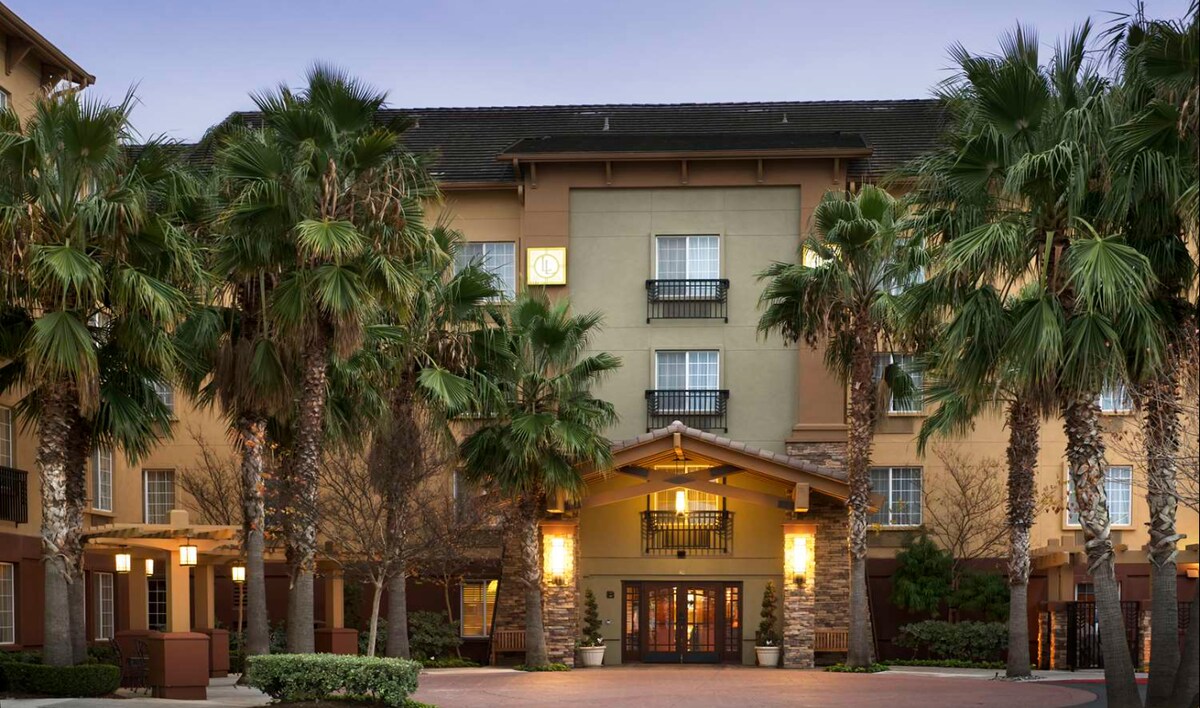 The hotel exterior features a welcoming entrance framed by tall palm trees. A covered entryway leads to large glass doors, providing a glimpse into the lobby. The building's contemporary design is complemented by warm, inviting lighting along the façade.