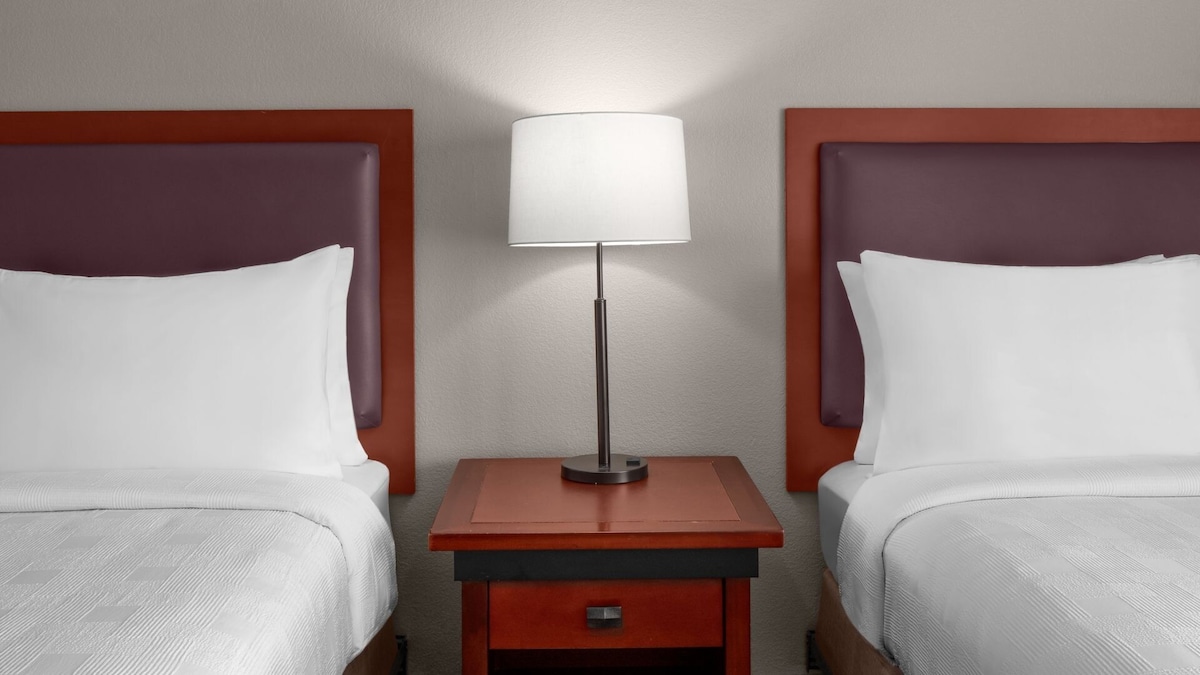 A hotel room featuring two Queen beds with soft white linens and flat, upholstered headboards in a muted purple. A nightstand with a modern lamp rests between the beds, providing a warm, diffused light against the neutral wall.