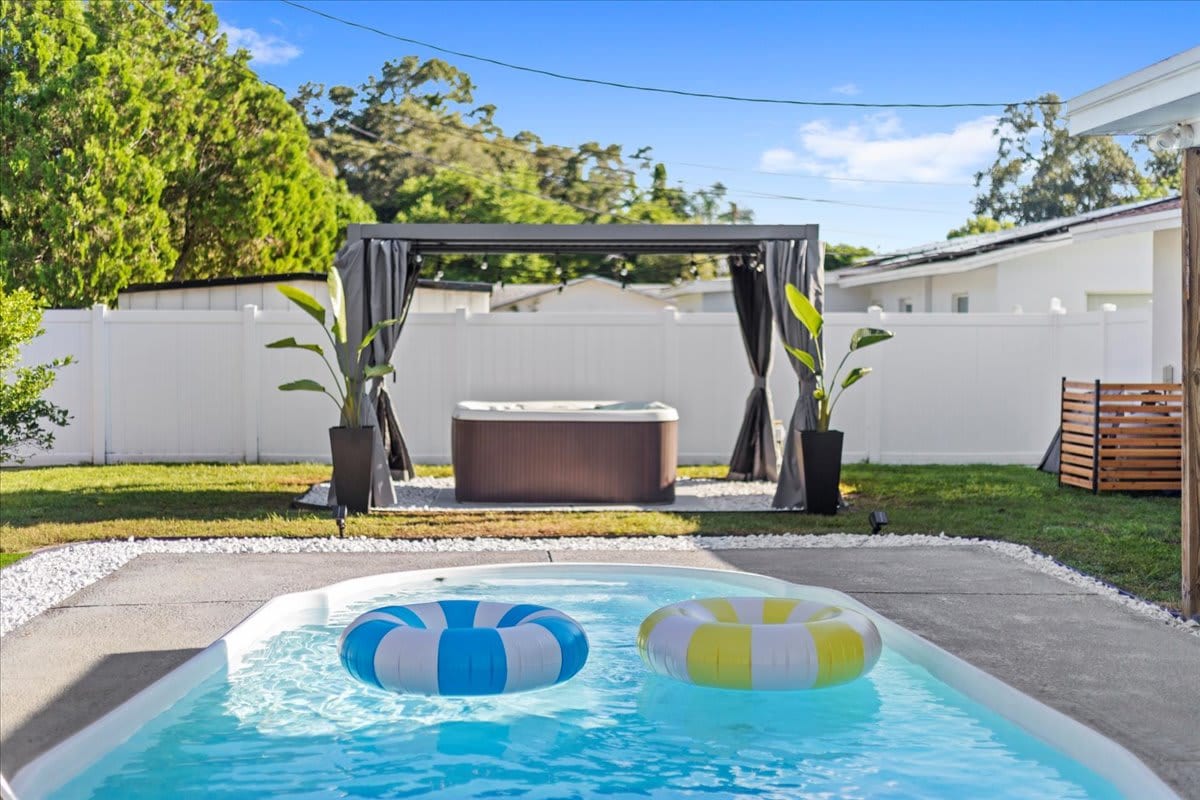 A sparkling pool is framed by a paved patio, featuring two colorful inflatable rings floating on the surface. A covered hot tub, surrounded by greenery and curtains, is positioned in the background, offering a relaxing outdoor retreat.