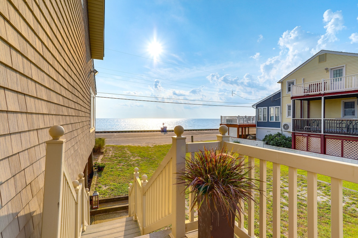 A staircase leads downwards from a beige house towards a grassy area and the bay, with the sun shining brightly in a blue sky. In the distance, boats can be seen on the water, and neighboring houses are visible on either side.
