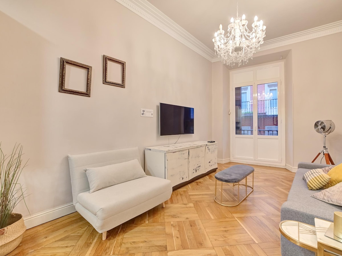The living area features a light-colored sofa bed and a comfortable armchair. A modern television is mounted on the wall above a stylish cabinet. Elegant crown molding frames the space, and natural light enters through large doors leading to a balcony.