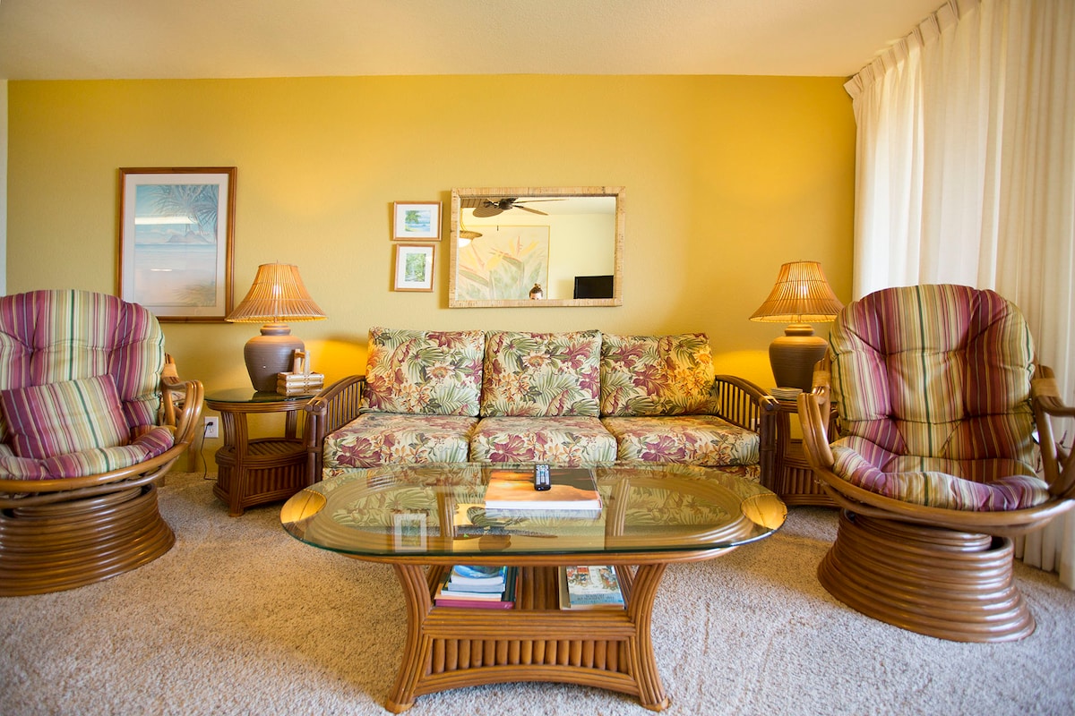 A cozy living area showcases a floral-patterned sofa, complemented by two swivel chairs in a striped fabric. A glass coffee table sits in front, featuring a stack of magazines and a remote. The warm yellow walls enhance the welcoming ambiance, while soft lighting is provided by two table lamps.