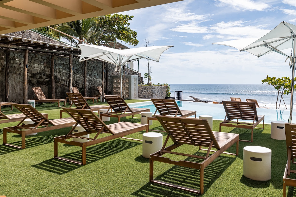 Lounge chairs arranged on a green grassy area overlook the ocean. A poolside bar structure and umbrellas provide shade nearby. The serene water stretches out towards the horizon, with a few palm trees and beachside elements subtly framing the view.