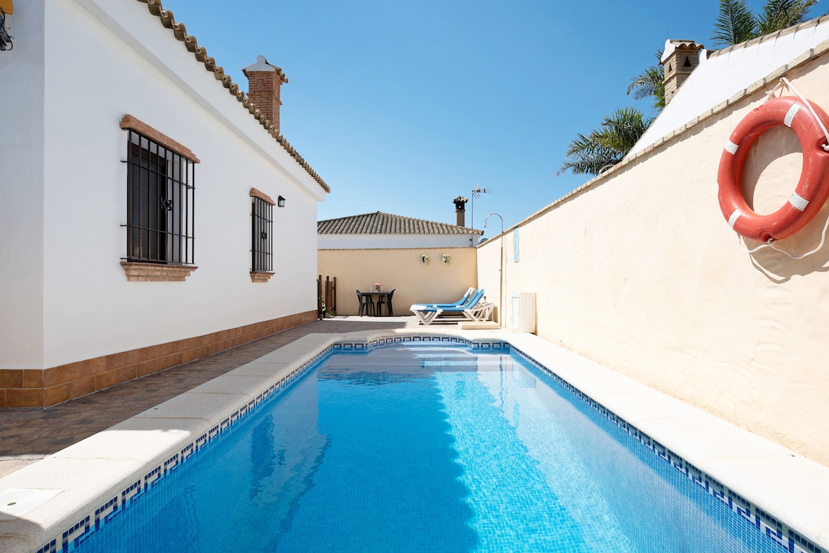 A private pool is surrounded by a sunlit patio. The pool features clean, blue water reflecting the sky, with a pair of sun loungers positioned nearby. A life preserver hangs on the wall, and a shaded seating area is visible in the background.