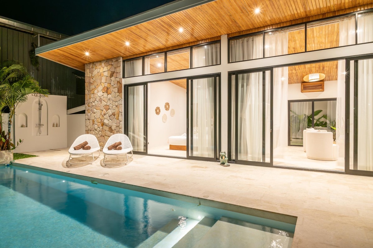 An inviting outdoor space features a serene blue swimming pool alongside a spacious patio. Large glass doors open to a bright interior, showcasing natural wood accents and modern furnishings. Comfortable seating is arranged near the pool, blending indoor and outdoor living areas.