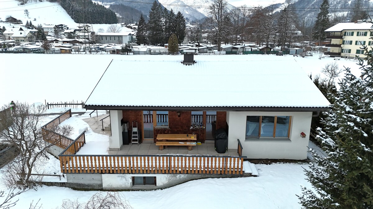 The exterior of a cozy holiday home is shown, surrounded by snow-covered ground and trees. A spacious deck extends from the house, featuring a wooden table and benches for outdoor seating. The roof is snow-capped, and the nearby mountains create a serene backdrop.