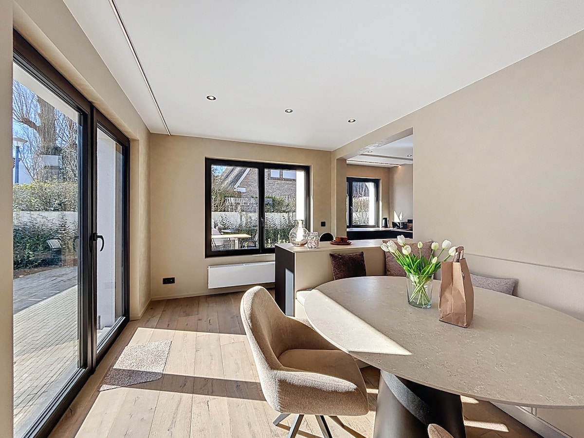 The dining area is furnished with a round table surrounded by beige chairs, illuminated by natural light from large glass doors. Soft colors and simple decor create a serene atmosphere, with a vase of white tulips as a focal point on the table.