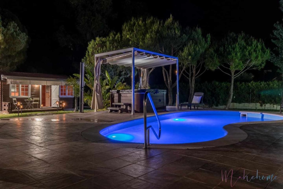 A tranquil scene is presented at night, featuring a softly illuminated swimming pool with blue lighting. Nearby, a covered seating area offers comfort beside a hot tub, surrounded by lush trees and a charming bungalow that adds to the serene atmosphere.