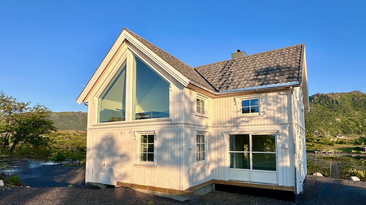 A modern house with a triangular glass feature stands under a clear blue sky. Soft natural light is reflected on the white wooden exterior. The structure showcases a mix of traditional and contemporary design elements, surrounded by greenery and scenic mountain views.