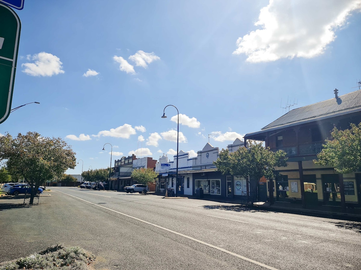 A quiet street is lined with historic buildings featuring a mix of shopfronts and residential facades. A clear blue sky and bright sunlight enhance the scenery, with trees providing scattered shade along the roadside.