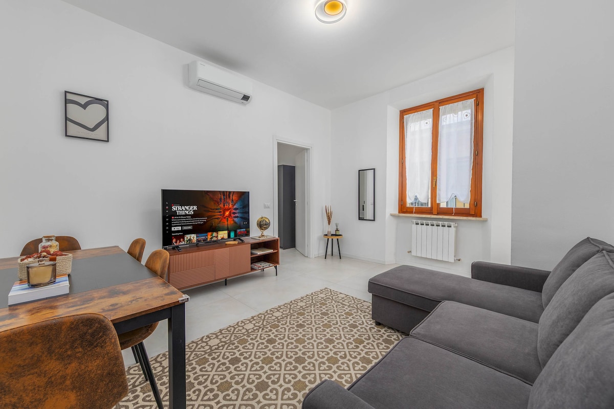 A bright living room is showcased, featuring a dark sofa and a wooden dining table with four chairs. A smart TV is positioned on a stylish cabinet. Natural light enters through a large window, illuminating the space. A decorative rug adds warmth to the floor.