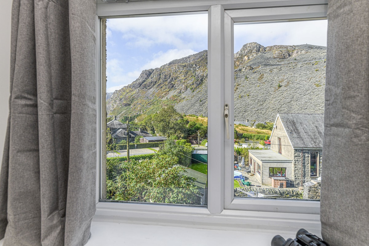 A window frames the stunning view of rugged hills and mountainous terrain. Lush greenery is visible below, along with traditional buildings that blend into the landscape. Light grey curtains softly drape on either side, enhancing the natural light entering the space.