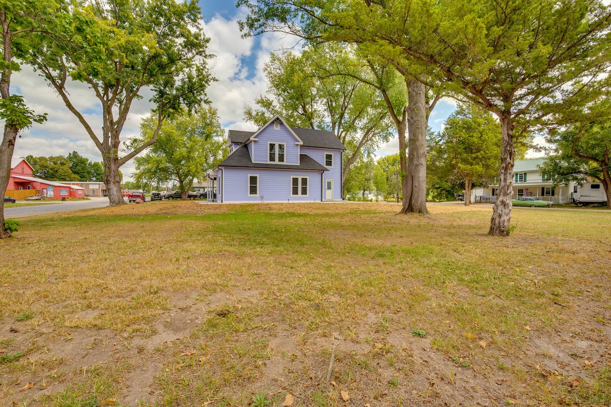 A two-story purple house is surrounded by grassy grounds and mature trees. The property offers a spacious outdoor area, with ample sunlight filtering through the leaves. Nearby structures and a roadway are visible in the distance.