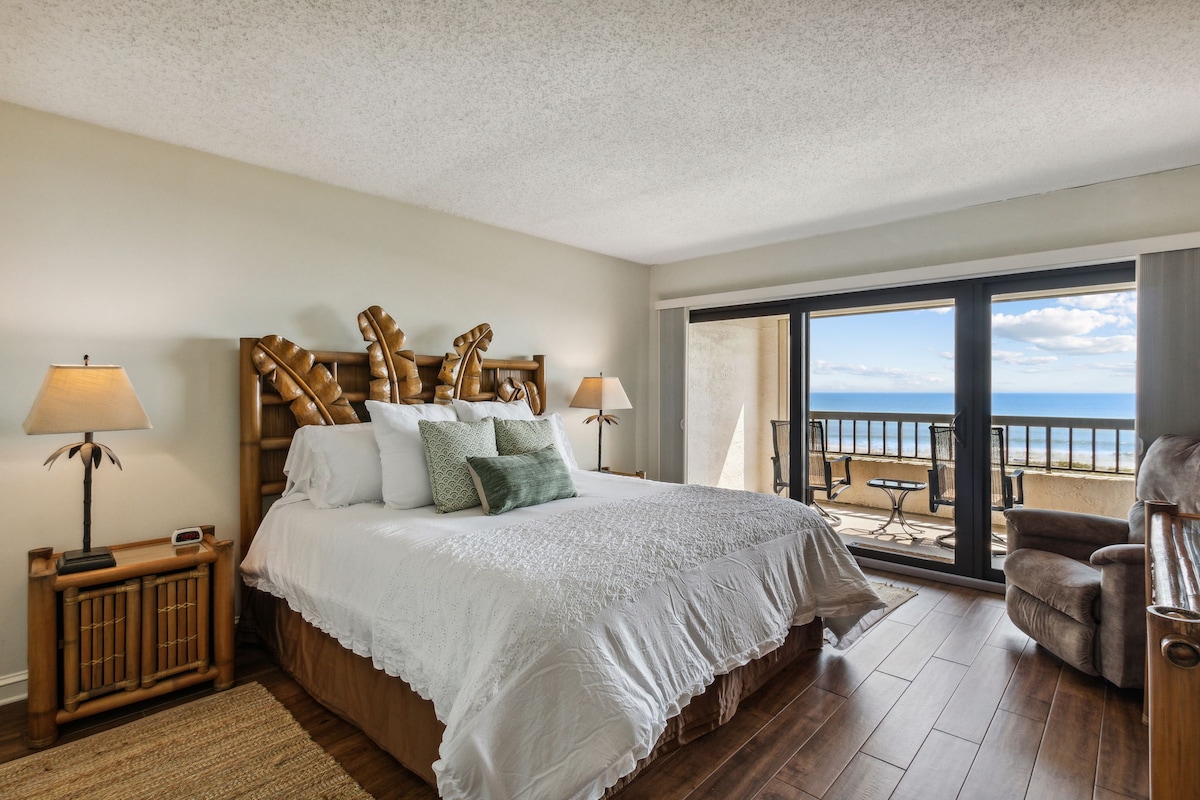 The bedroom features a spacious bed dressed in crisp white linens, with decorative pillows adding a touch of color. Natural light floods the room through large sliding glass doors that open to a private balcony, revealing views of the ocean and outdoor seating.