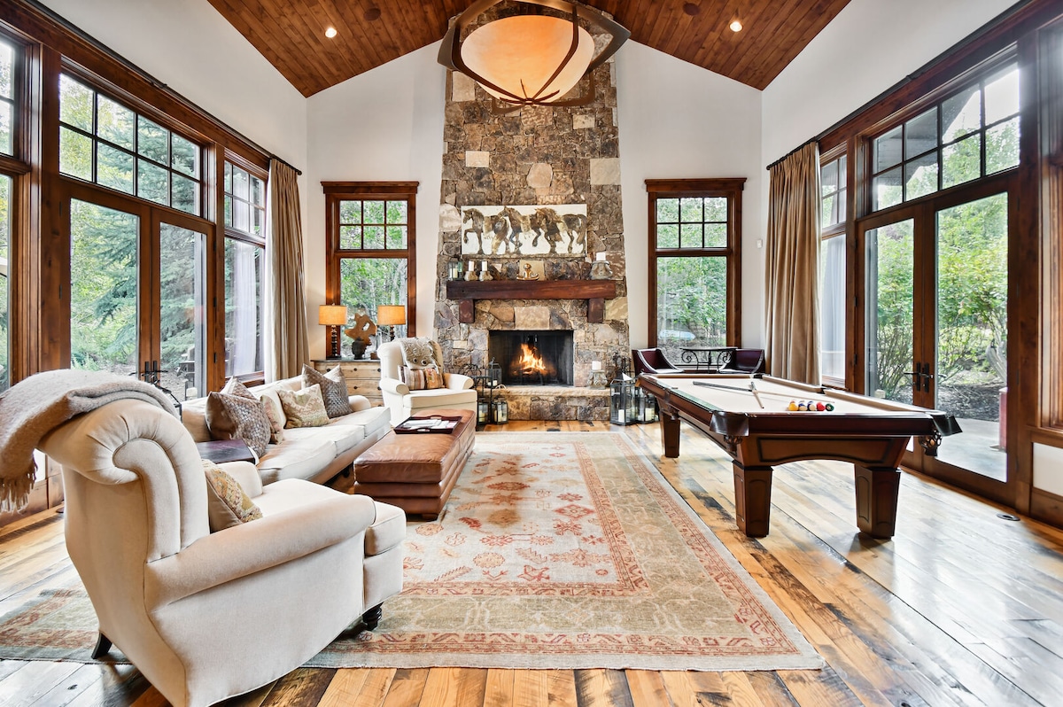 A spacious living area features a grand stone fireplace, elegant wood beams, and large windows that offer views of the outdoors. A comfortable white sofa and neutral armchair are arranged around a decorative area rug, while a pool table provides an inviting entertainment option.