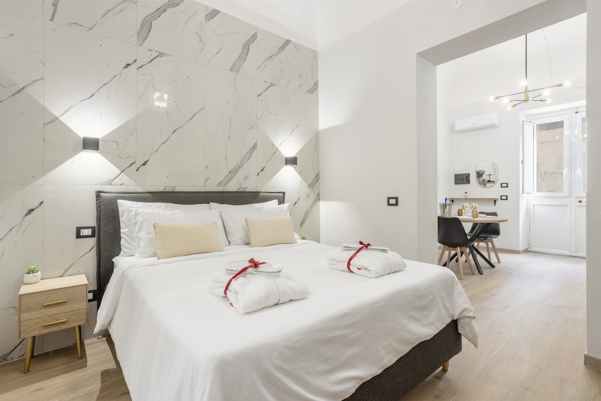 A spacious bedroom is observed, featuring a large bed with neatly arranged white linens and decorative towels. Neutral tones are complemented by marble accent walls. In the background, a dining area is visible, showcasing a table and chairs near a window that allows natural light.