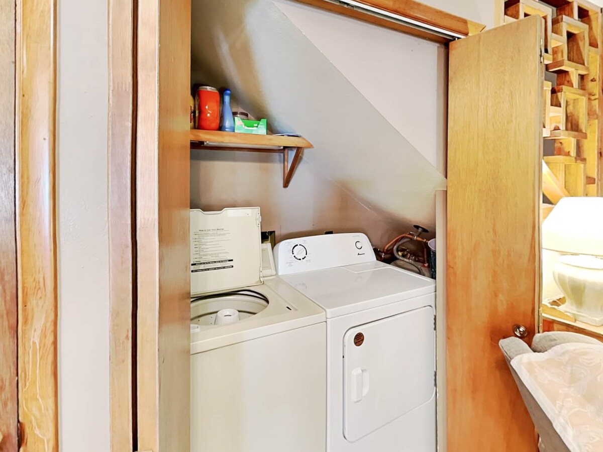 A utility space is shown housing a stacked washer and dryer. Shelves above hold a few cleaning supplies and laundry items. Soft lighting from a nearby lamp provides a warm glow to the area, highlighting the wood features of the closet's interior.