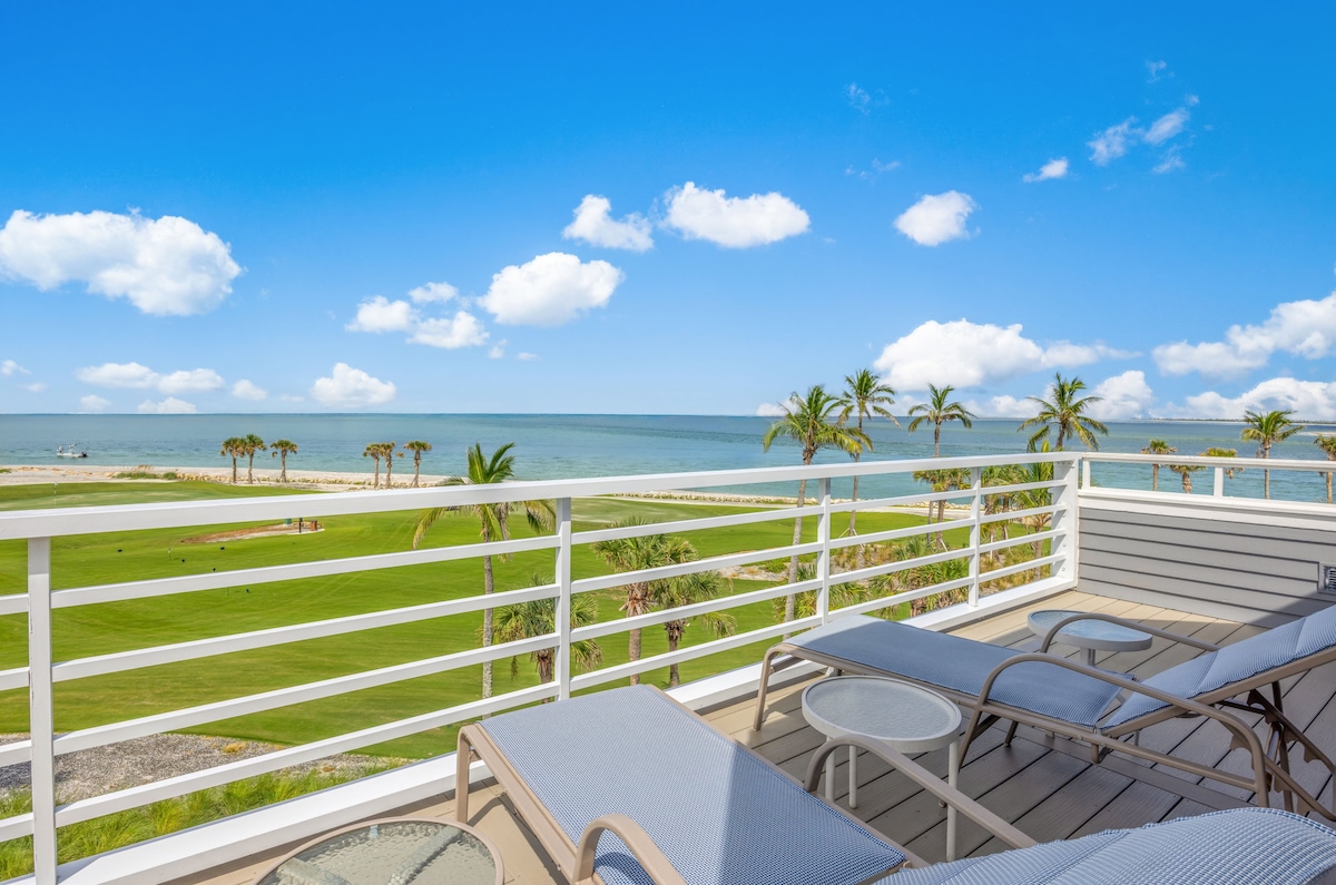A spacious balcony overlooks the Gulf, framed by swaying palm trees and lush green grass. Comfortable lounge chairs are positioned for optimal views of the serene waters and distant horizon, complemented by a clear blue sky scattered with fluffy white clouds.