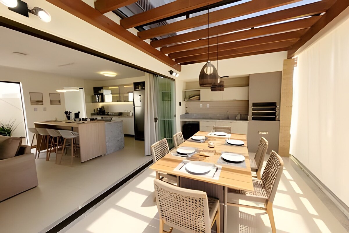 An open dining area features a wooden table set for meals, surrounded by woven chairs. Large sliding doors provide a view of a modern kitchen, brightened by natural light from overhead beams. The space is complemented by minimalist decor and neutral colors.