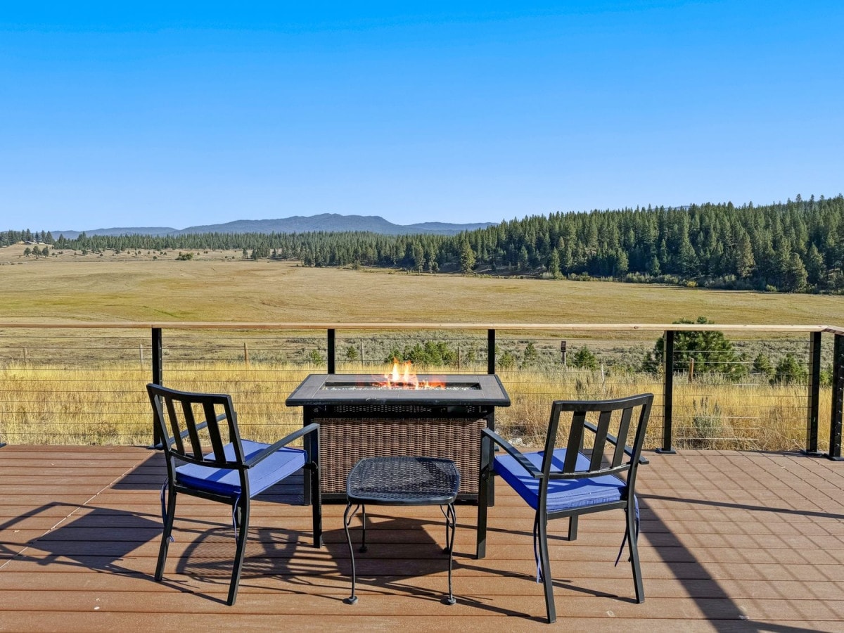 A deck is featured with two black chairs positioned around a propane fire table, creating a cozy gathering space. Expansive views of rolling hills and distant mountains are visible in the background, under a clear blue sky.