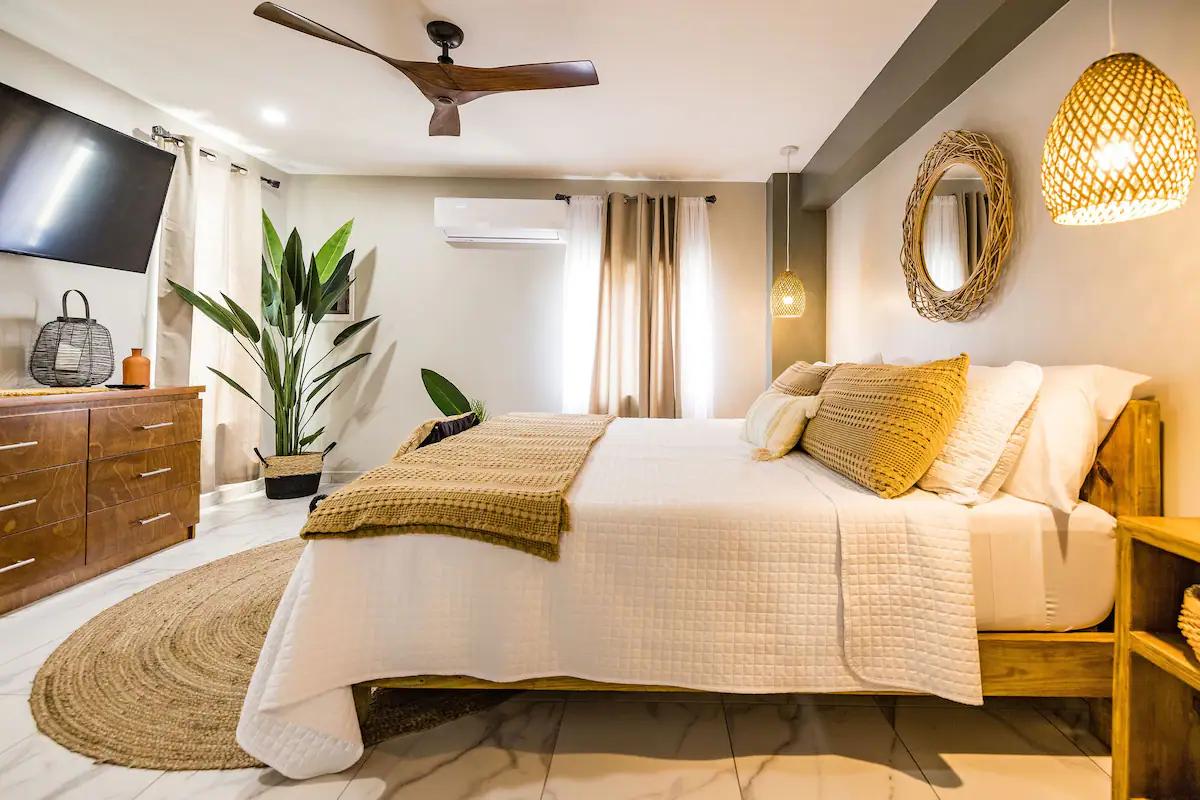 A cozy bedroom is furnished with a spacious bed adorned with a textured blanket and decorative pillows. A calming color palette is complemented by natural light filtering through sheer curtains. Plant accents and woven decor enhance the inviting ambiance of this serene retreat.