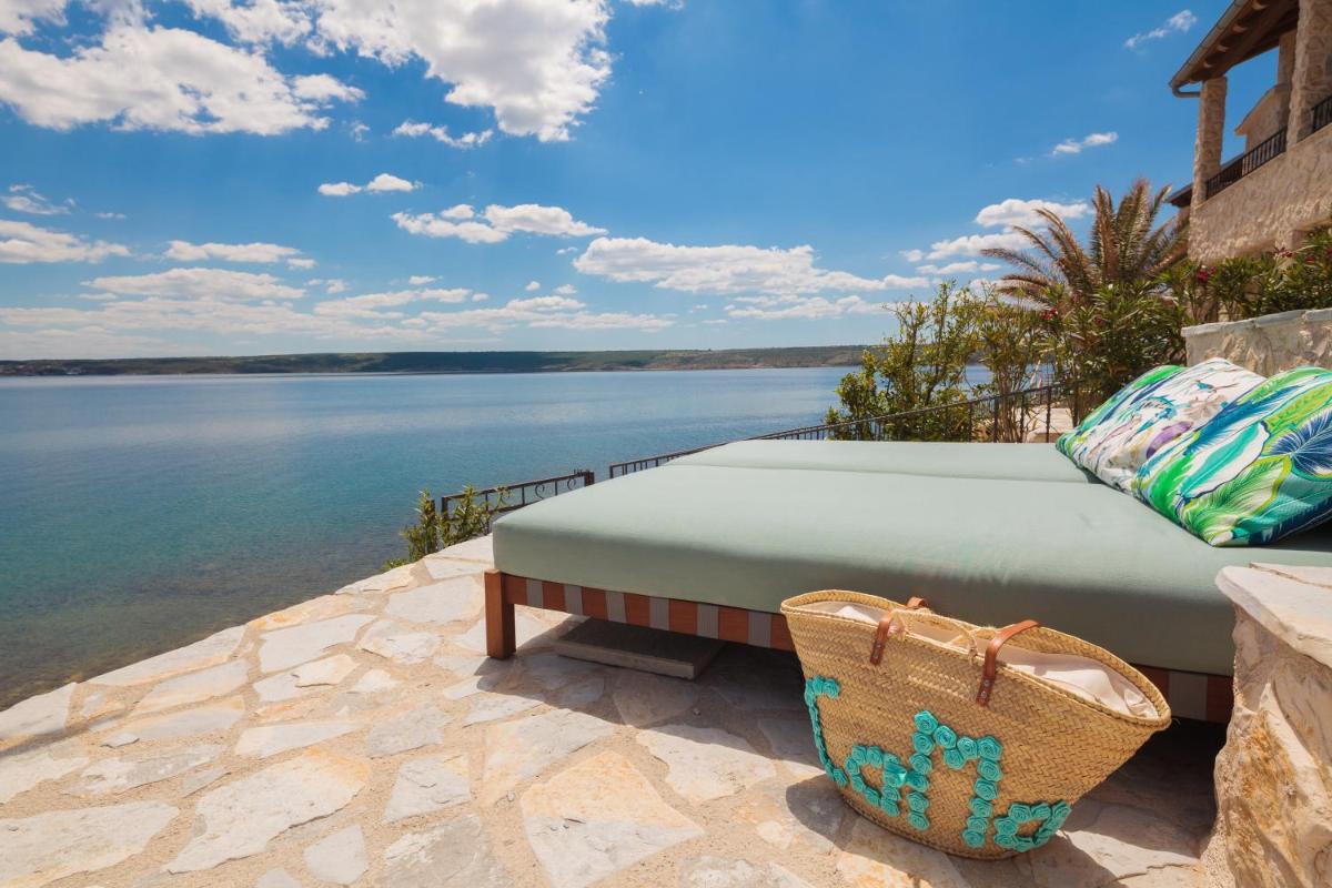 A sun lounger with decorative cushions rests on a stone terrace, overlooking calm waters under a bright sky dotted with clouds. A woven beach bag sits nearby, adding a touch of charm to the serene coastal scene.
