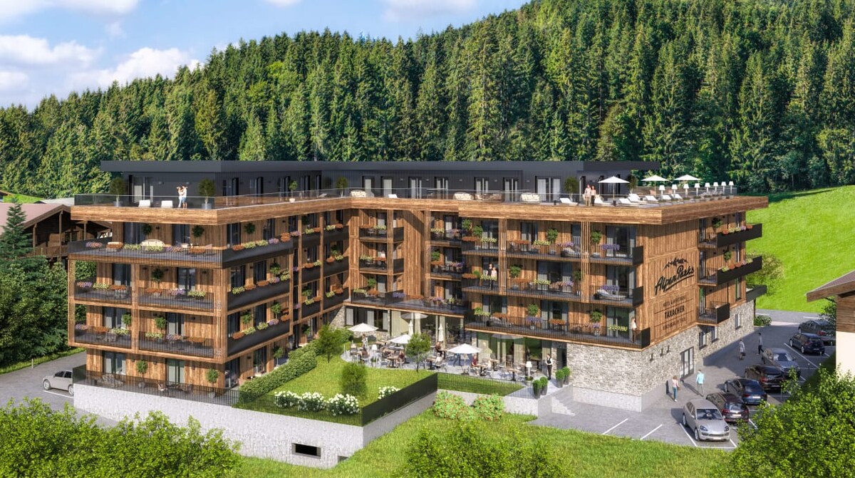 The modern hotel building features a wooden facade with multiple balconies. Lush green trees provide a natural backdrop, while the structured layout showcases outdoor seating areas and terraces, promoting a connection with the surrounding landscape.