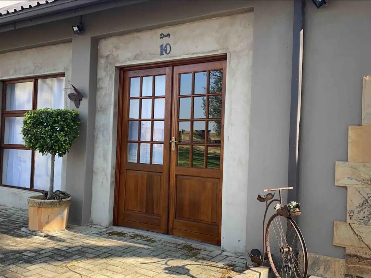 The entrance features double wooden doors with glass panes, framed by two windows. A small topiary plant in a pot adds greenery beside the door, while a vintage bicycle stands against the building's exterior, complementing the rustic charm of the area.