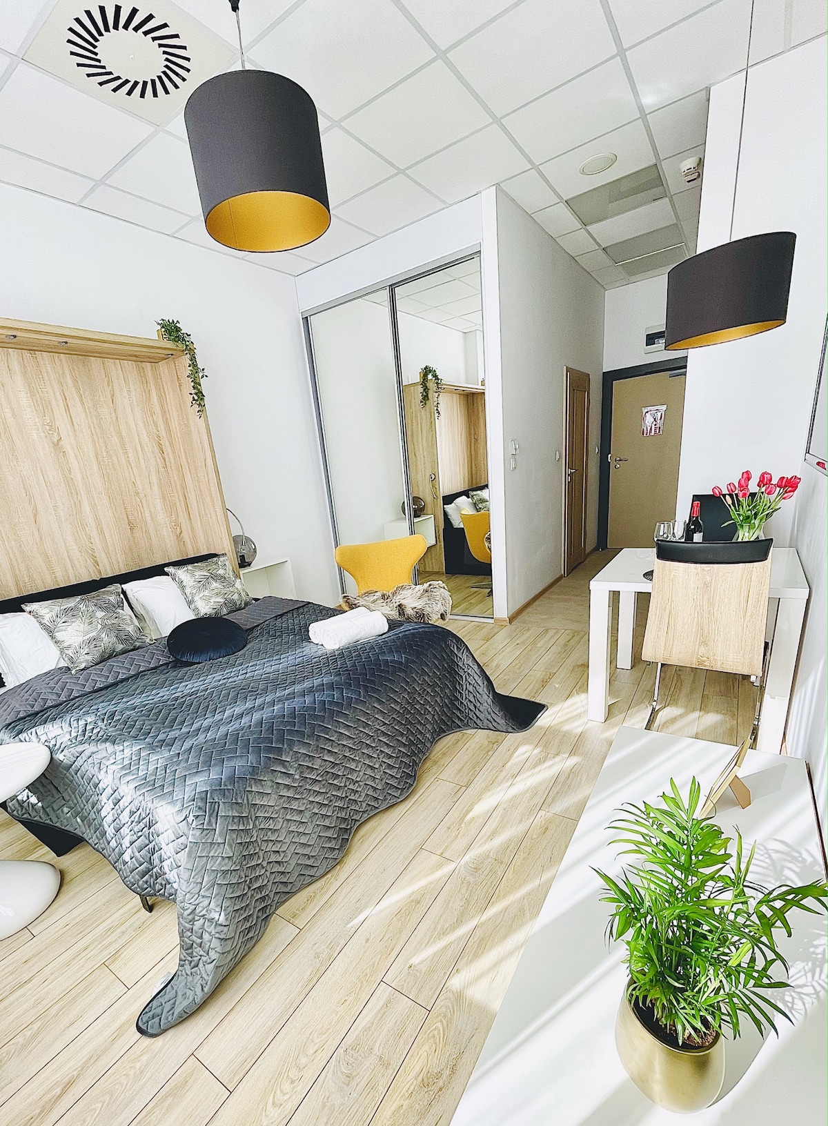 The room features a double bed with a textured gray blanket and decorative pillows. A small table with a plant sits next to the bed. A sitting area with a yellow chair is visible, and mirrors reflect the space, enhancing the brightness.