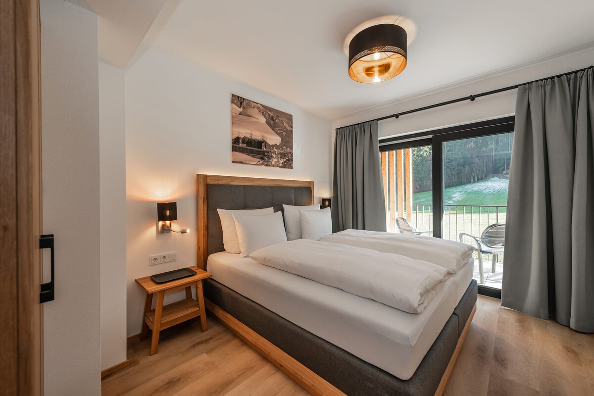 A comfortable master bedroom features a spacious bed with crisp linens. Two bedside lamps provide soft lighting, and large glass doors offer access to an outdoor balcony. Soft gray curtains frame the windows, enhancing a calm and restful ambiance.