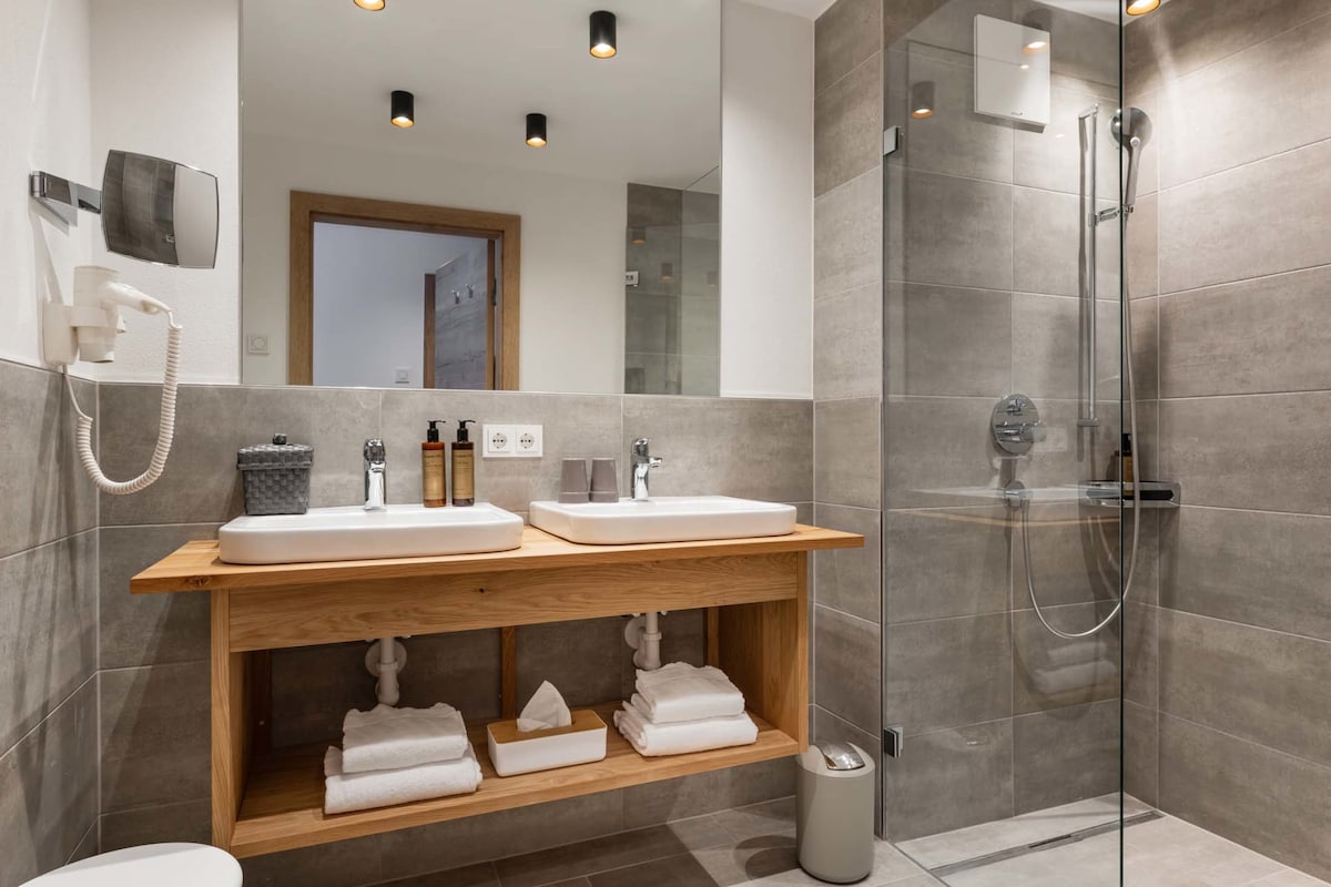The bathroom features a modern design with two under-mounted sinks set on a wooden countertop. A spacious walk-in shower is enclosed by clear glass. White towels are neatly arranged on a shelf beneath the sinks, complemented by toiletries displayed on the countertop.