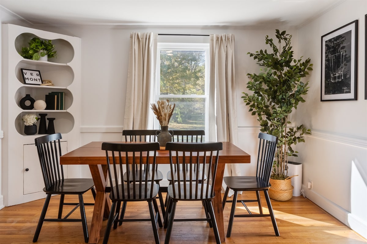 A dining area is furnished with a wooden table surrounded by six black chairs. Natural light enters through a window, complemented by sheer curtains. A potted plant and decorative items are displayed on a shelf, adding to the room's inviting atmosphere.