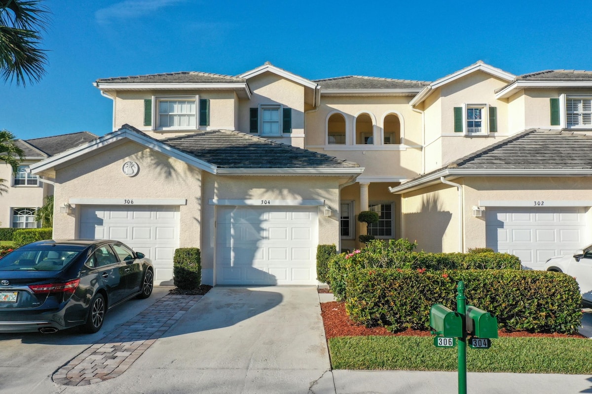 The exterior of a two-story townhome complex is displayed, featuring a light-colored façade with decorative arched windows. Two garages are visible, along with manicured landscaping that includes shrubs and palm trees. A blue sky and bright sunlight illuminate the scene.