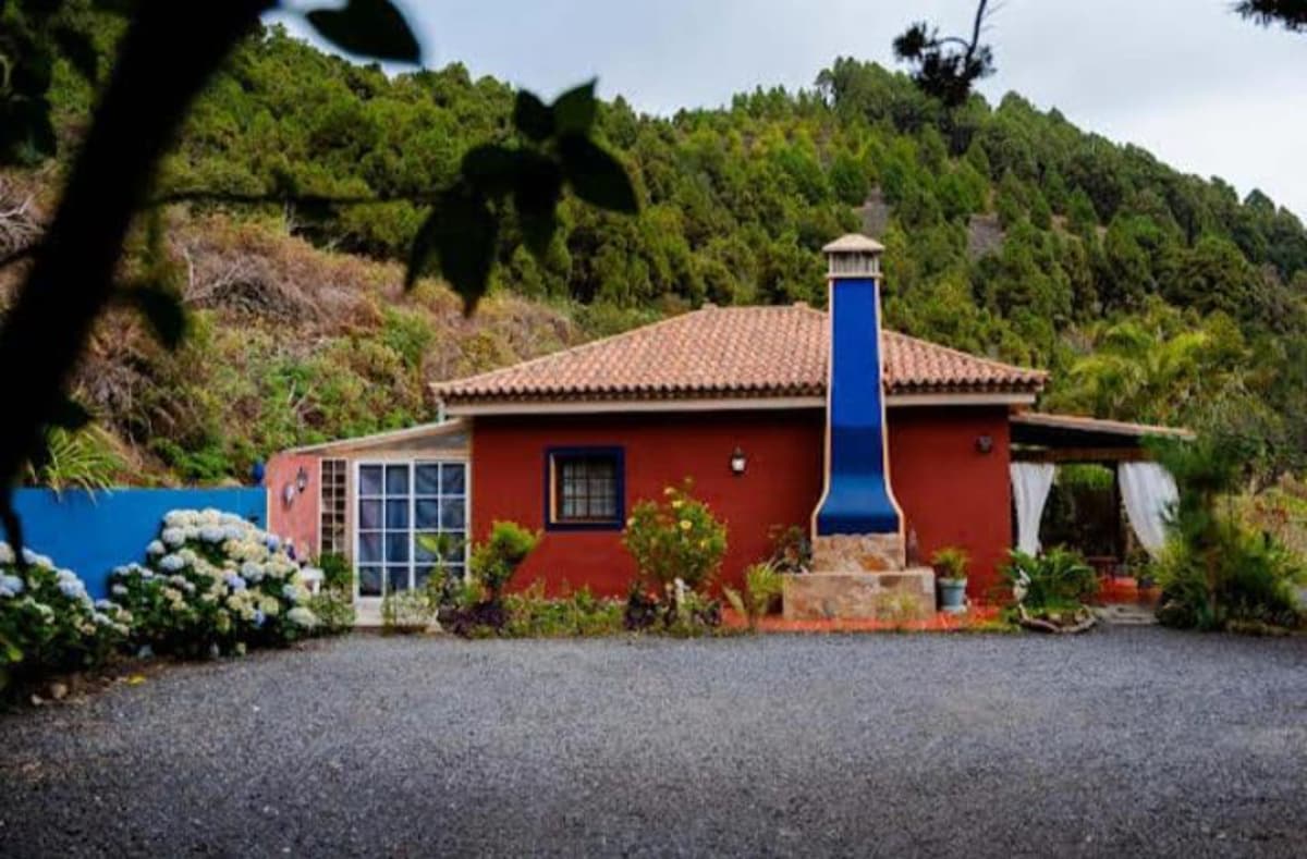 The exterior of the holiday apartment is showcased, featuring a vibrant red facade complemented by blue accents and a distinctive chimney. A gravel driveway leads to the entrance, surrounded by lush greenery and flowering shrubs. The rustic roof adds character to the overall appearance.