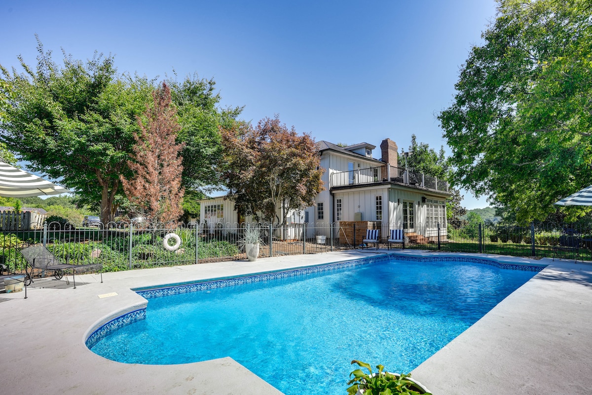 A private pool is surrounded by well-maintained landscaping, featuring lush trees and vibrant plants. Comfortable seating is arranged nearby, providing space for relaxation. The main cottage is visible in the background, complemented by a blue sky overhead.