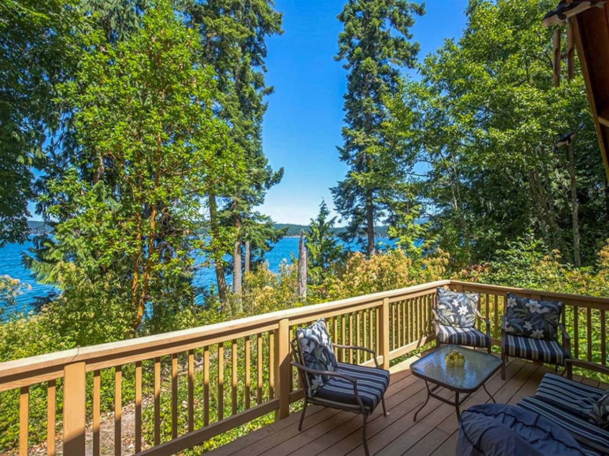 The deck offers a serene view of Sequim Bay framed by lush greenery. Comfortable seating is arranged around a glass-topped table, inviting relaxation in the warm sunlight. The surrounding trees provide a sense of privacy while capturing glimpses of the water through the foliage.