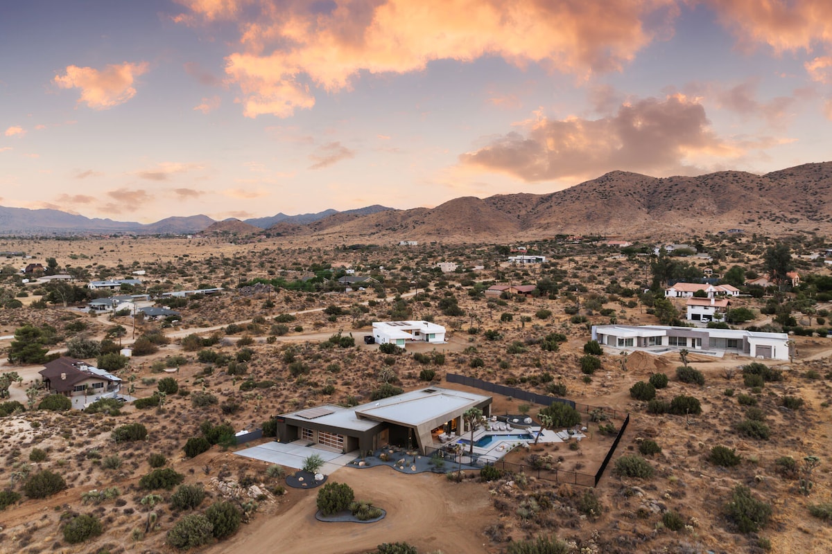 An aerial view captures the stylish villa nestled in the desert landscape, surrounded by rugged hills and sparse vegetation. A stunning sunset casts warm hues across the sky, illuminating the villa's sleek architecture and inviting outdoor spaces.