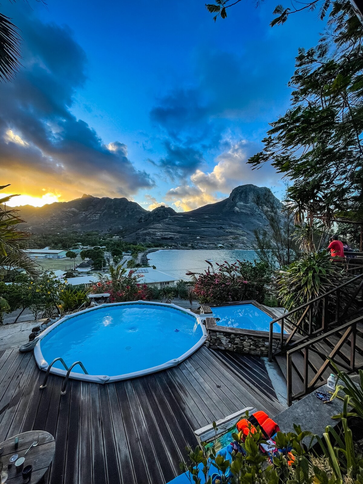 Two inviting hot tubs are positioned on a wooden deck, surrounded by lush foliage. Mountain peaks rise in the background, with a serene bay and village visible below. The sky is painted with warm hues from the setting sun, creating a tranquil atmosphere.