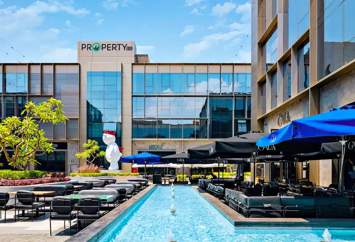 An outdoor area features a sleek swimming pool bordered by modern black lounge chairs. Tall glass structures reflect the blue sky, while a vibrant tree adds greenery to the space. Umbrellas provide shade over the seating area, creating a relaxing atmosphere.
