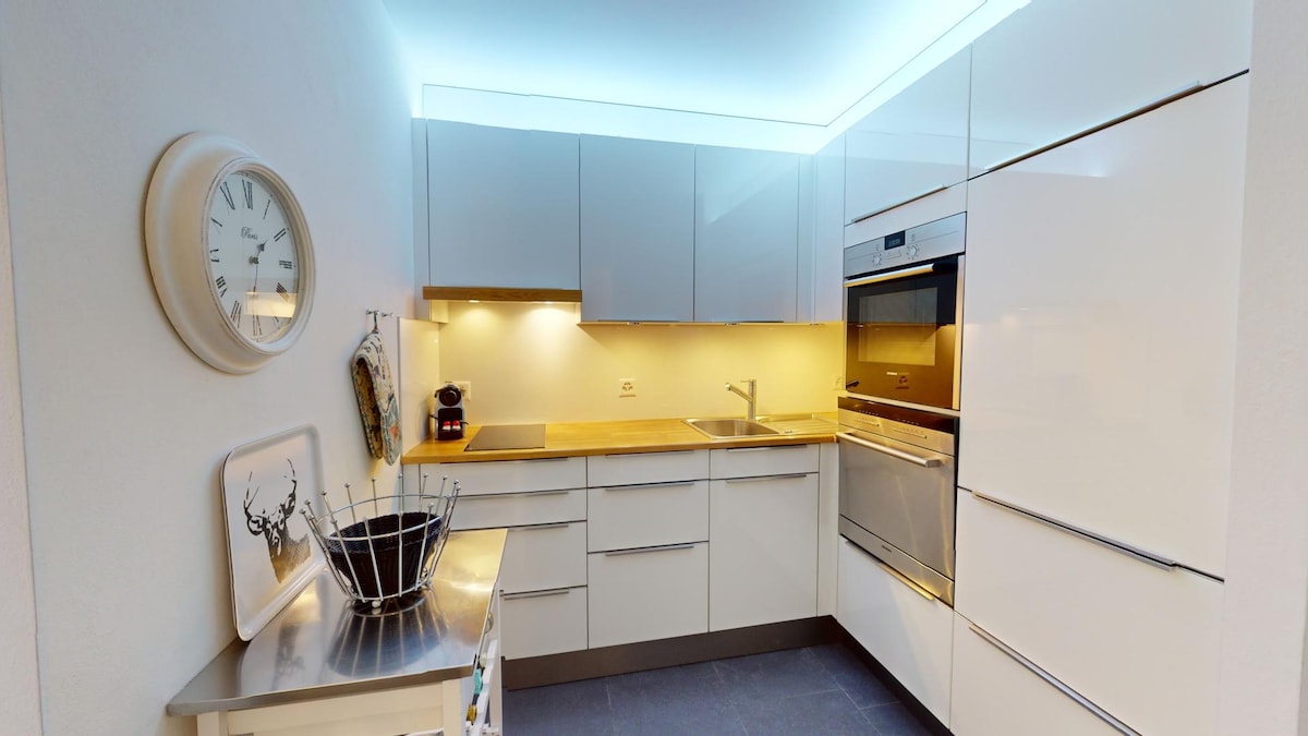 A modern kitchen features sleek, white cabinetry and optimized storage. A wooden countertop complements stainless steel appliances, including an oven and coffee maker. Soft under-cabinet lighting provides visibility, while a wall clock adds functionality.