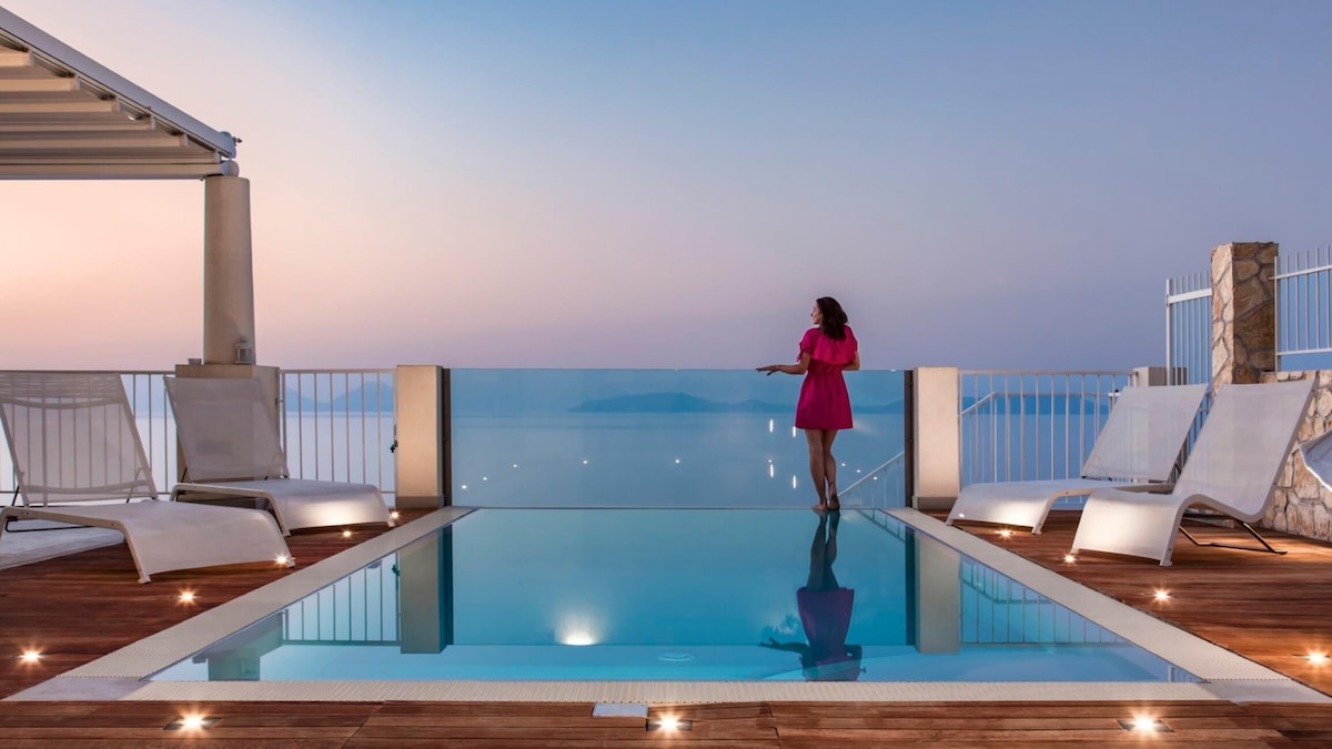 An infinity pool offers stunning views of the sea at dusk. The area is framed by sleek lounge chairs and soft lighting, creating a serene environment. The horizon is visible in the background, enhancing the peaceful atmosphere.
