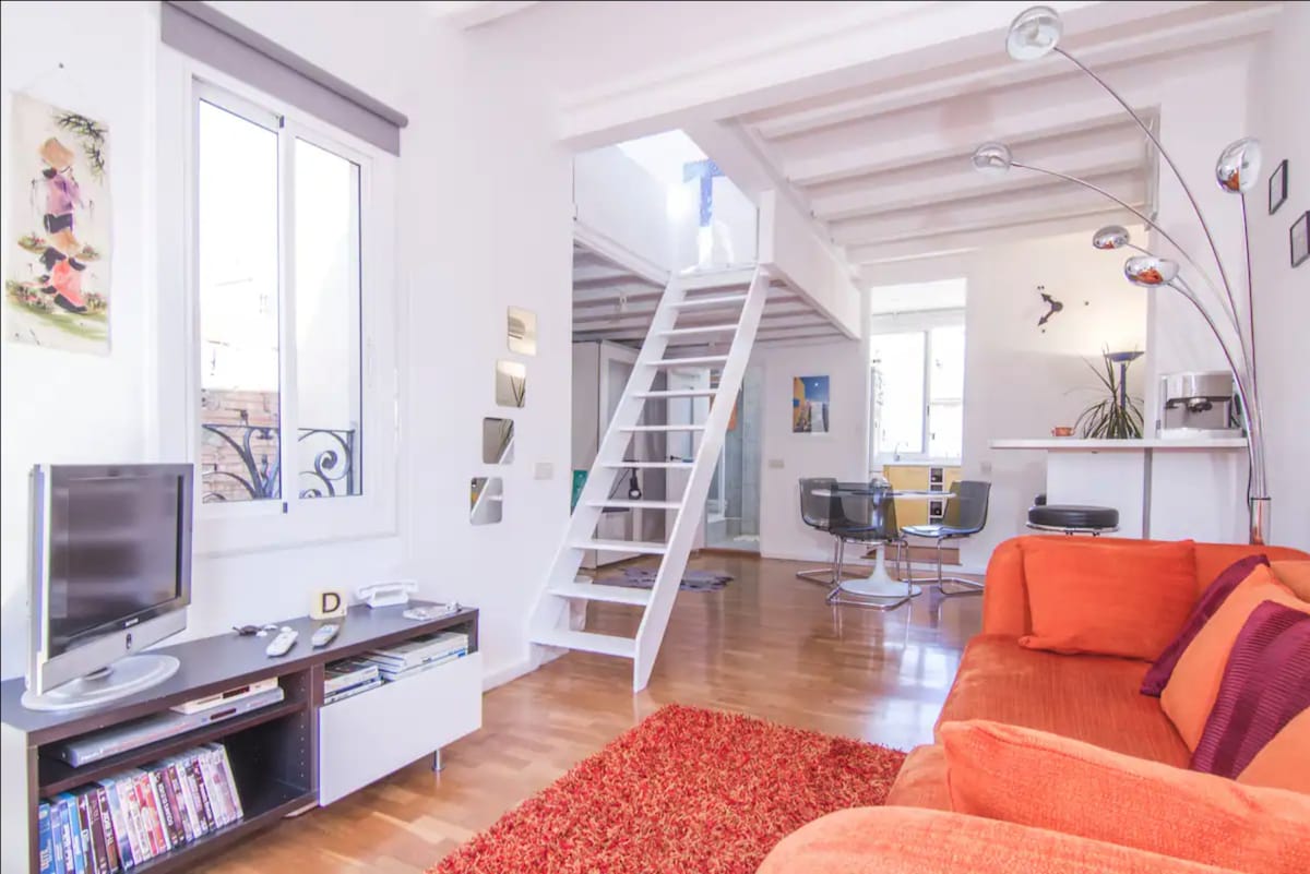 An open living area features a bright orange sofa, complemented by a textured red rug. A modern TV unit is situated beside the sofa, while sleek dining chairs surround a glass table in the background. A staircase leads to the upper level, enhancing the loft's spaciousness.