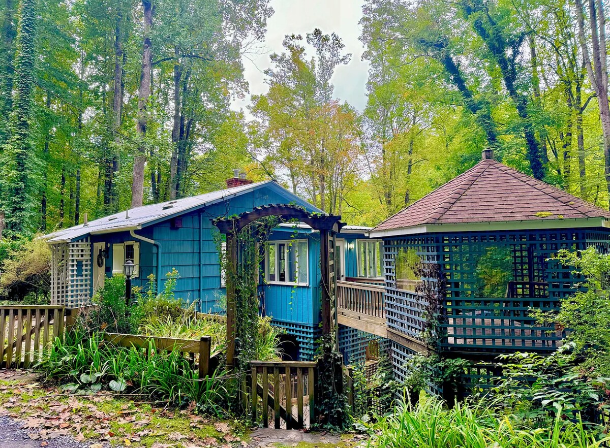 A charming blue mountain cabin is surrounded by tall trees and vibrant foliage. An inviting gazebo is positioned nearby, showcasing a wooden lattice design. The structure features a bridge connecting to the main cabin, enhancing the serene ambiance of the outdoor space.