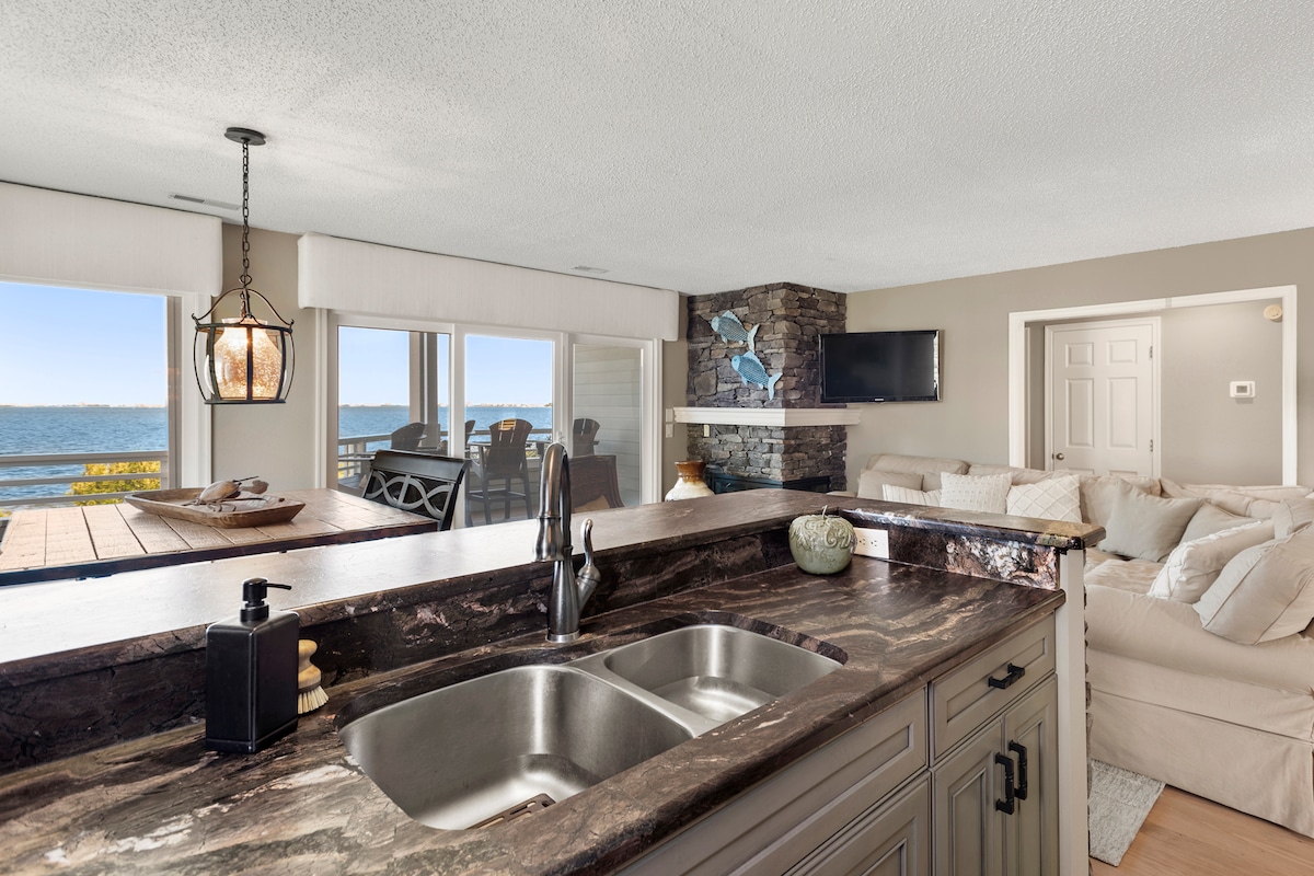 The open kitchen displays sleek dark countertops and a modern double sink. A view of the water is visible through expansive windows, while a cozy living area is seen in the background with comfortable seating and decorative elements.